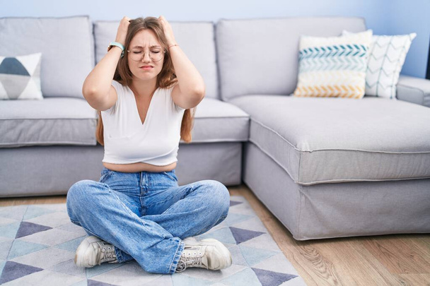 Молодая белая женщина, сидящая на полу в гостиной, страдает от головной боли в отчаянии и стрессе из-за боли и мигрени. руки на голове.  - Фото, изображение