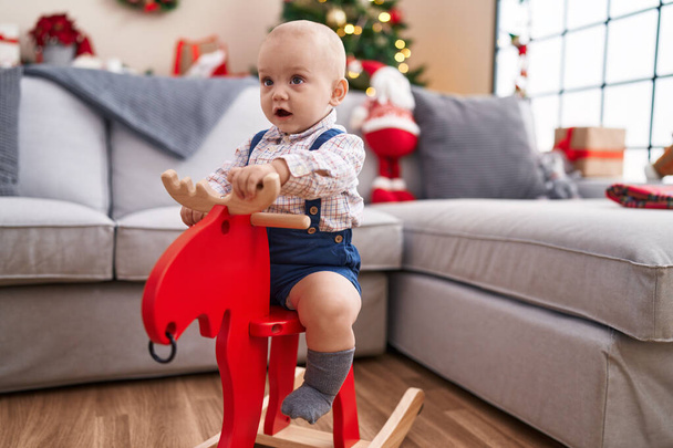Suloinen valkoihoinen vauva leikkii poron kanssa keinuen joulukuusen luona kotona - Valokuva, kuva