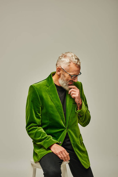handsome cheerful mature man in vibrant attire with glasses and beard sitting on tall chair - Photo, Image