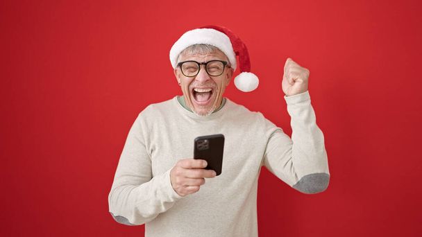 Grauhaariger Mann mittleren Alters trägt Weihnachtsmütze mit Smartphone über isoliertem rotem Hintergrund - Foto, Bild
