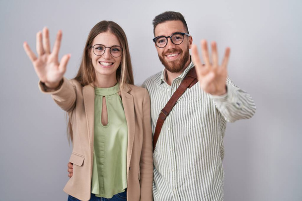 Junges Paar steht vor weißem Hintergrund und zeigt mit Finger Nummer neun nach oben, während es selbstbewusst und glücklich lächelt.  - Foto, Bild