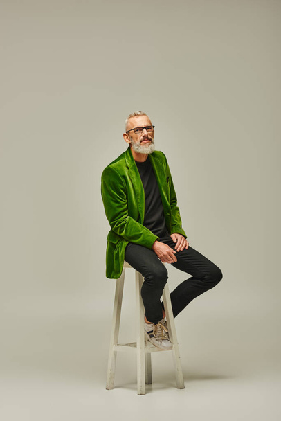 good looking mature male model with beard and glasses sitting on tall chair and looking at camera - Photo, Image