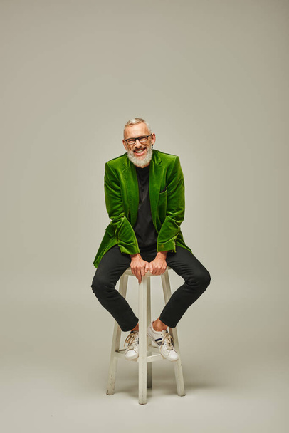 alegre modelo masculino funky com barba e óculos sentados em cadeira alta e sorrindo para a câmera - Foto, Imagem