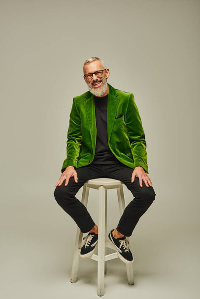 homem maduro atraente barbudo alegre em blazer verde sentado em cadeira alta e sorrindo para a câmera - Foto, Imagem