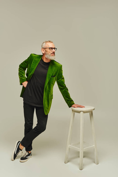 good looking mature man with beard in vibrant attire posing next to tall chair and looking away - Photo, Image