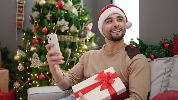 Young arab man using smartphone holding gift sitting by christmas tree at home - Photo, Image