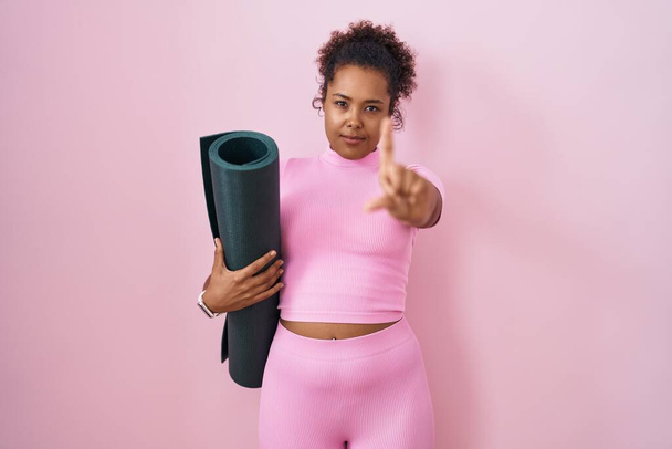 Молодая латиноамериканка с вьющимися волосами, держащая коврик для йоги на розовом фоне, показывая пальцем вверх и сердитым выражением лица, не показывая жеста  - Фото, изображение