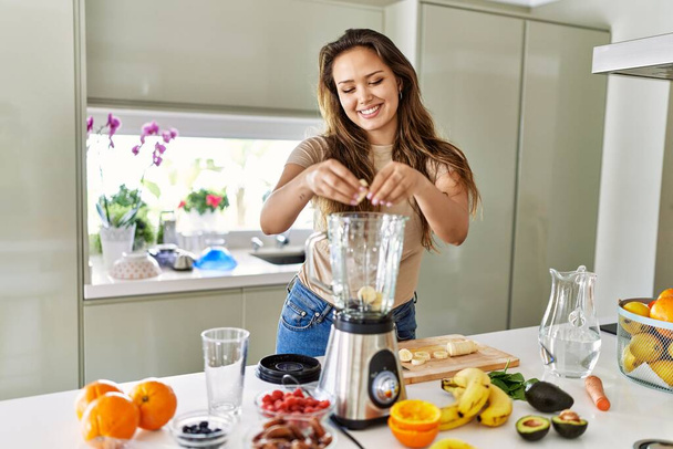 Jovem bela mulher hispânica preparando smoothie vegetal com liquidificador na cozinha - Foto, Imagem