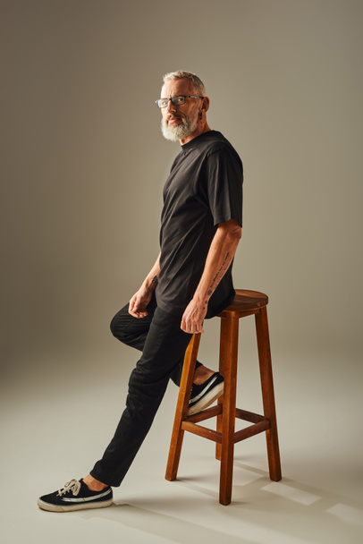 knap volwassen man in zwart t shirt zitten op lange stoel en kijken naar camera op beige achtergrond - Foto, afbeelding