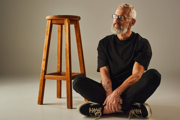 atraente maduro alegre homem no preto t-shirt com tatuagens sentado no chão com pernas cruzadas - Foto, Imagem