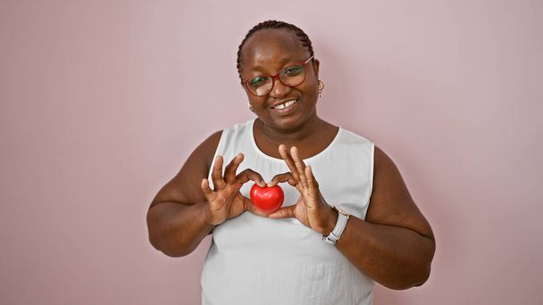A magabiztos afro-amerikai nő örömmel tartja a szív alakját a kezével, mosolyog az elszigetelt rózsaszín fal háttér, kifejező pozitív életmód - Fotó, kép
