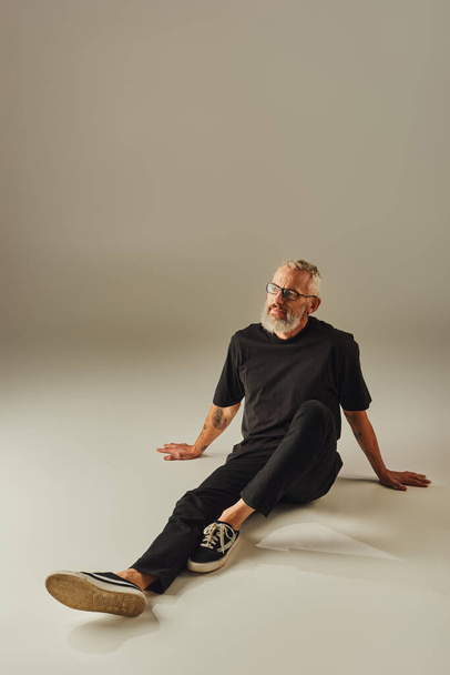 vrolijk volwassen man in zwart t shirt met bril zitten op de vloer en weg te kijken op beige achtergrond - Foto, afbeelding