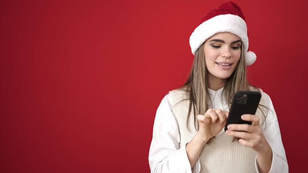 Giovane bella donna ispanica indossa cappello di Natale utilizzando smartphone su sfondo rosso isolato - Foto, immagini
