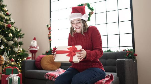jong blond vrouw uitpakken cadeau zitten op bank door kerstboom thuis - Foto, afbeelding