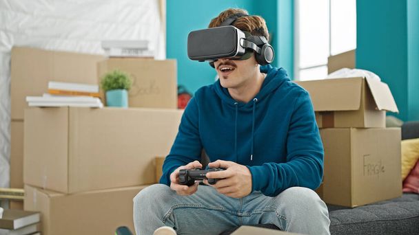 Jeune homme hispanique jouer à un jeu vidéo en utilisant joystick et lunettes de réalité virtuelle à la nouvelle maison - Photo, image