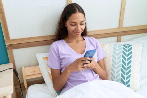 Young african american woman using smartphone sitting on bed at bedroom - Photo, Image