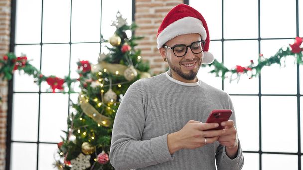 Hombre hispano usando sombrero de Navidad usando smartphone en casa - Foto, imagen