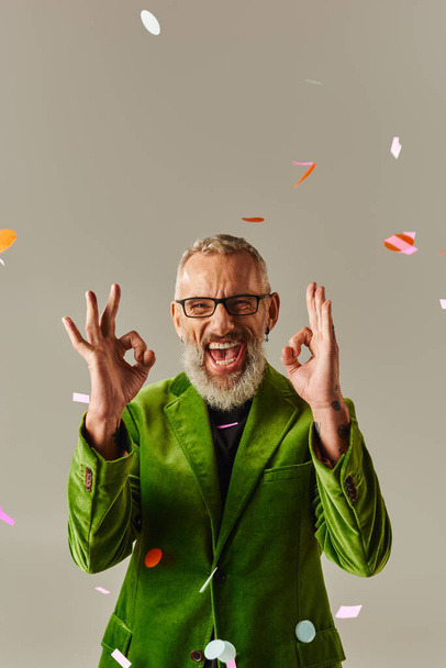 handsome jolly mature male model in green blazer showing okay sign at camera under confetti rain - Photo, Image