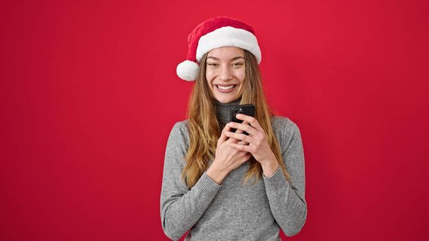 Mujer caucásica joven que usa sombrero de Navidad con teléfono inteligente sobre fondo rojo aislado - Foto, Imagen