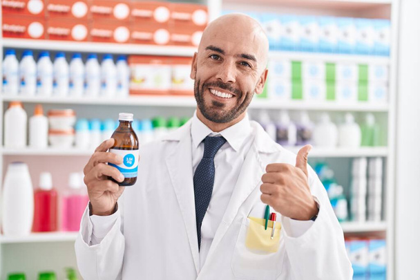 Homem careca de meia idade trabalhando na farmácia farmácia xarope segurando sorrindo feliz e positivo, polegar-se fazendo excelente e sinal de aprovação  - Foto, Imagem