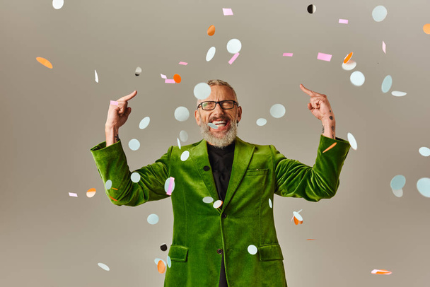 cheeky jolly mature man with closed eyes in green blazer showing middle fingers under confetti rain - Photo, Image
