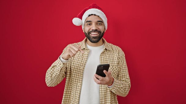Młody Hiszpan w świątecznym kapeluszu wskazuje na smartfona na odizolowanym czerwonym tle - Zdjęcie, obraz