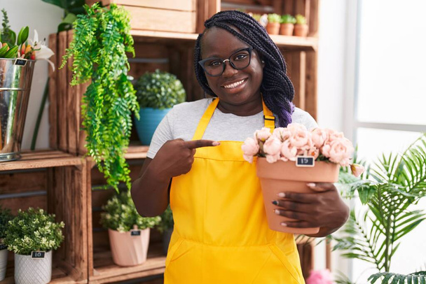 Молодая африканская женщина работает в цветочном магазине держа завод улыбаясь счастливо указывая рукой и пальцем  - Фото, изображение