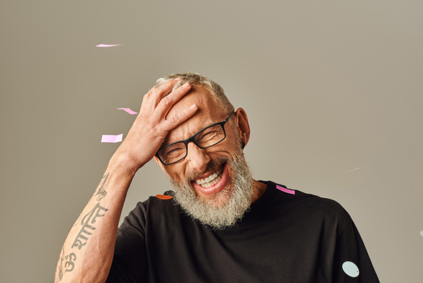 good looking jolly mature man in black t shirt with tattoo under confetti rain with hand on head - Fotoğraf, Görsel