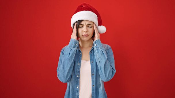Νεαρή λευκή γυναίκα που φοράει χριστουγεννιάτικο καπέλο και έχει πονοκέφαλο σε απομονωμένο κόκκινο φόντο - Φωτογραφία, εικόνα