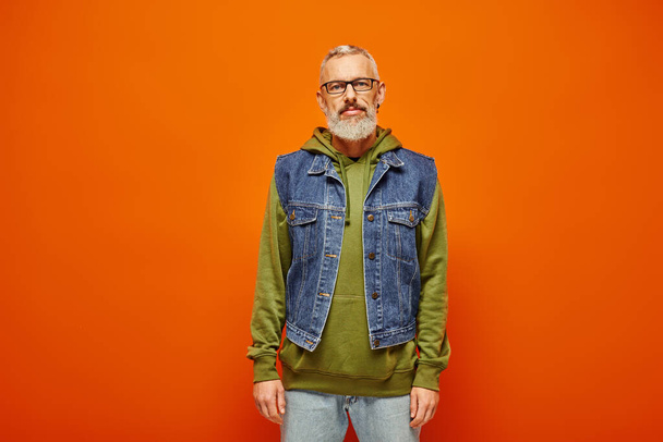 schöner, reifer Mann in grünem Kapuzenpulli und stylischer Jeansweste, der vor orangefarbenem Hintergrund in die Kamera blickt - Foto, Bild