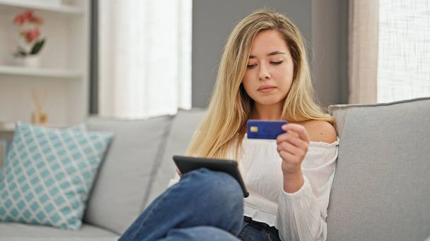 Młoda blondynka zakupy z touchpad i karty kredytowej siedzi na kanapie w domu - Zdjęcie, obraz