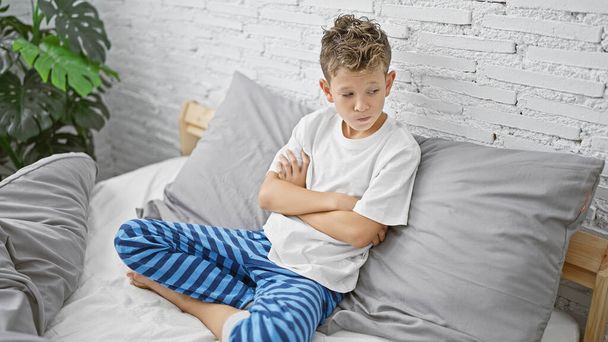 Adorable garçon blond, assis sur le lit avec les bras croisés, l'air bouleversé dans la chambre un triste scénario du matin. - Photo, image