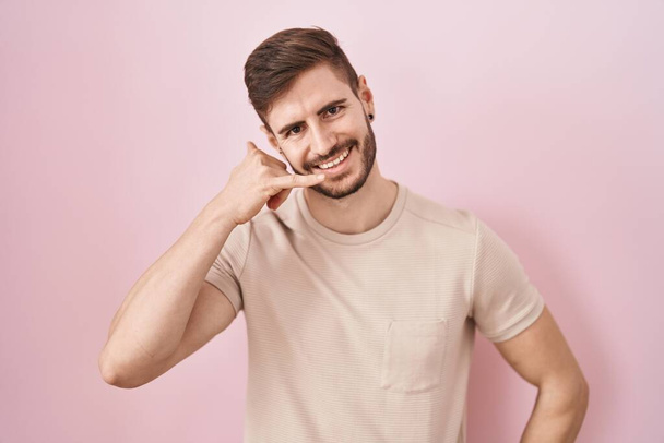 Латиноамериканец с бородой стоит на розовом фоне и улыбается, делая телефонный жест рукой и пальцами, как будто разговаривая по телефону. коммуникационные концепции.  - Фото, изображение