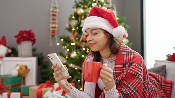 Νεαρή όμορφη ισπανόφωνη γυναίκα που χρησιμοποιεί smartphone κάθεται δίπλα στο χριστουγεννιάτικο δέντρο πίνοντας καφέ στο σπίτι - Φωτογραφία, εικόνα