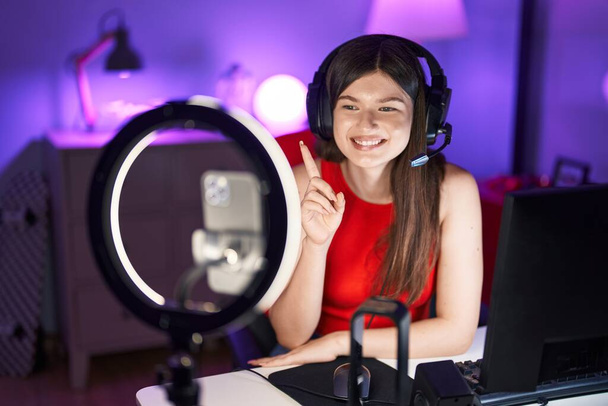 Jeune femme caucasienne jouant à des jeux vidéo enregistrement avec smartphone souriant heureux pointant avec la main et le doigt sur le côté  - Photo, image