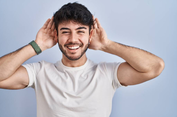 Hispanic man with beard standing over white background trying to hear both hands on ear gesture, curious for gossip. hearing problem, deaf  - 写真・画像