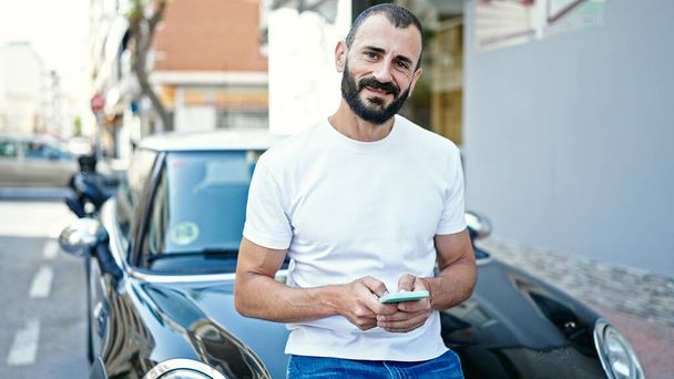 Young hispanic man using smartphone leaning on car at street - Foto, afbeelding