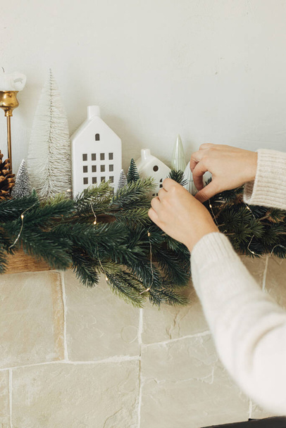 Decoratie van kerst open haard met verlichting, sparren takken, witte huisjes decoratie. Handen in gezellige trui opknoping decoratie op mantel in moderne boerderij. Sfeervolle wintervakantie - Foto, afbeelding