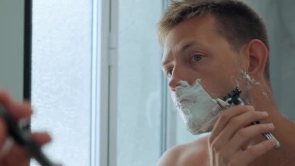 Hombre guapo yong caucásico con espuma de afeitar en su cara mientras se afeita la barba de pie en el baño y mirando al espejo. Rutina diaria de la mañana procedimiento de belleza higiénico masculino. - Metraje, vídeo