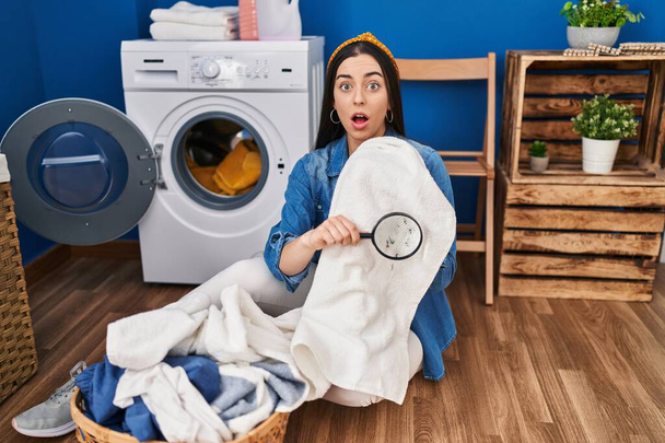 Hispanic woman holding magnifying glass looking for stain at clothes afraid and shocked with surprise and amazed expression, fear and excited face.  - Photo, image