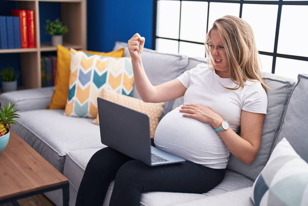 Junge schwangere Frau sitzt zu Hause mit Laptop genervt und frustriert auf dem Sofa und schreit vor Wut, schreit verrückt vor Wut und hebt die Hand  - Foto, Bild