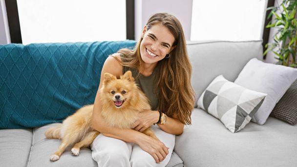 Mujer hispana joven confiada disfrutando de una risa divertida, abrazando a su perro mascota mientras está sentada alegremente en su sofá casero con una hermosa sonrisa. - Foto, imagen