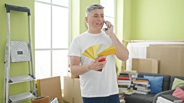 Νεαρός καυκάσιος άνδρας κρατώντας τεστ χρώματος μιλώντας στο smartphone στο νέο σπίτι - Φωτογραφία, εικόνα