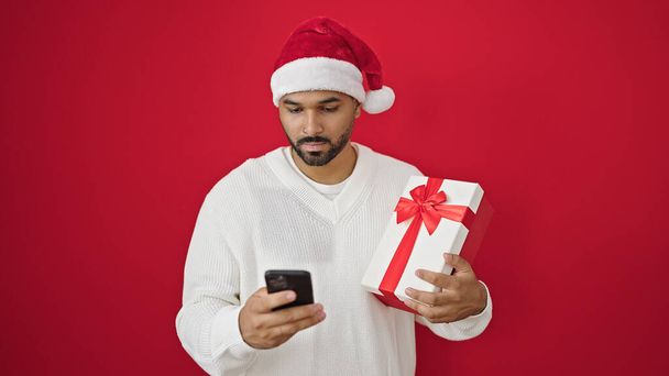 アフリカ系アメリカ人男性,隔離された赤い背景の上にスマートフォンを使用してクリスマスプレゼントを保持 - 写真・画像