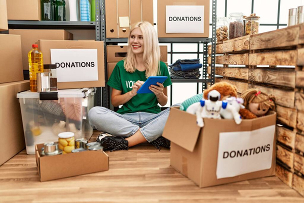 Молодая блондинка-волонтер использует тачпад, сидя на полу в благотворительном центре - Фото, изображение