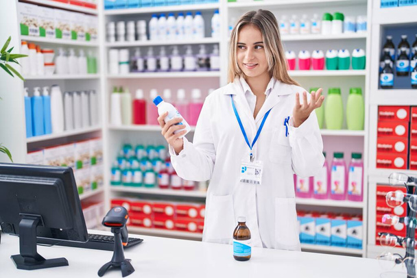 Joven mujer rubia farmacéutica sosteniendo frascos de medicamentos hablando en farmacia - Foto, imagen