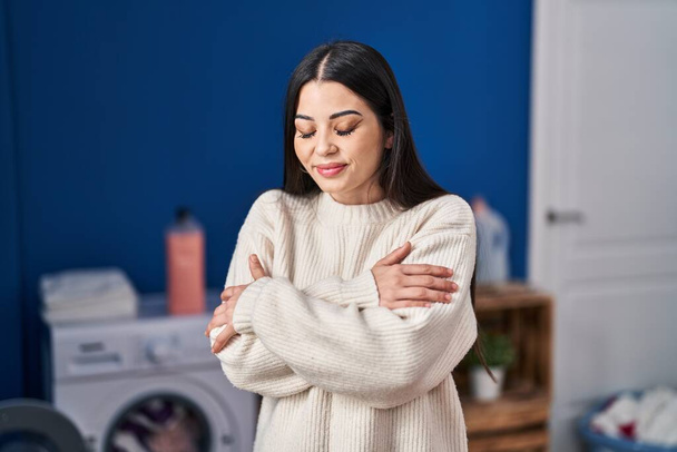 Young beautiful hispanic woman hugging himself touching softener sweater at laundry room - 写真・画像