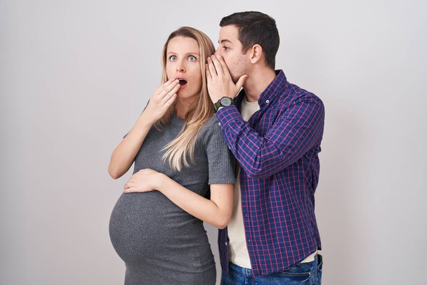 Jeune couple s'attendant à un bébé debout sur fond blanc main sur bouche racontant rumeur secrète, chuchotant conversation malicieuse  - Photo, image