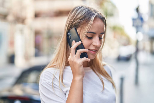 Молодая блондинка улыбается уверенно разговаривая по смартфону на улице - Фото, изображение
