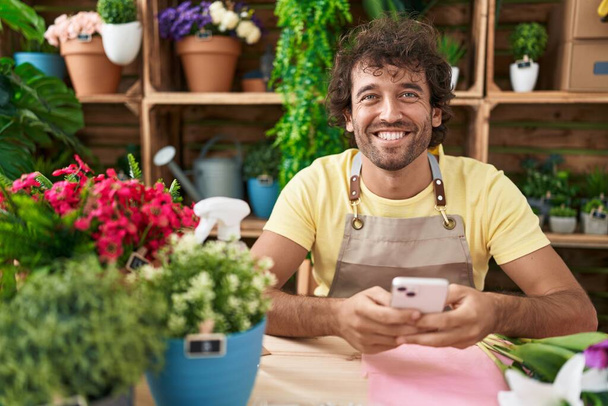 Молодий іспаномовний чоловік флорист посміхається впевнено, використовуючи смартфон у квітковому магазині
 - Фото, зображення